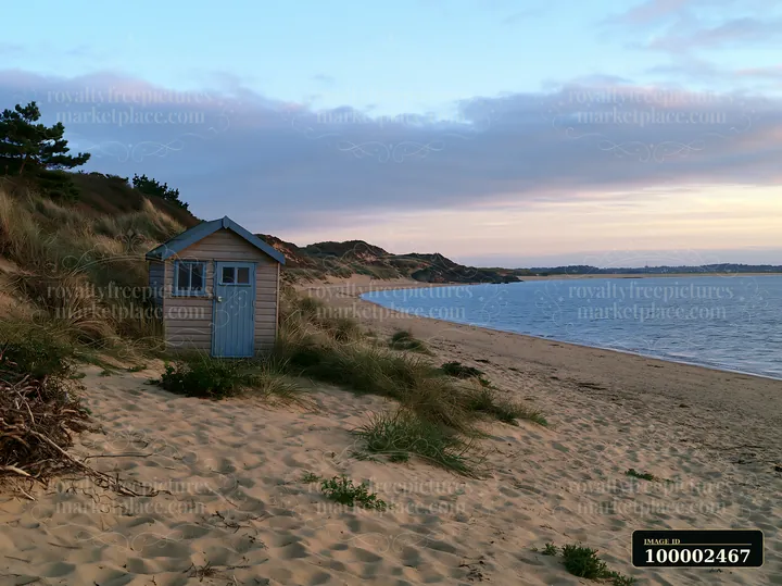 A small beach hut sits along the coastline with sheltered bay, surrounded by sand dunes and vegetation.
