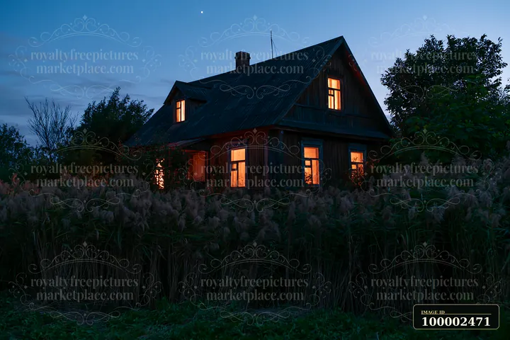 A wooden house is surrounded by tall grass with warm lights shining from the windows during the blue hour.