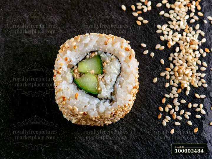 A single sushi piece sits on a black slate surface surrounded by scattered sesame seeds.