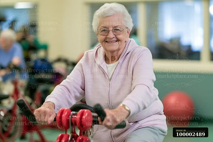 A senior with Parkinson's using a stationary bicycle smiles while exercising in a gym.