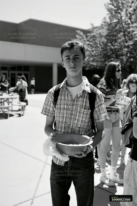 A young boy stands with a bowl in a line at a community potluck buffet during a sunny day outside a school.