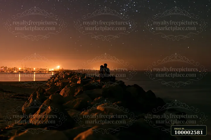 A couple shares a kiss silhouetted against a starry sky while seated on rocks at the beach.