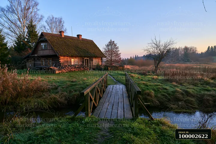 A rustic wooden cottage sits beside a small stream surrounded by fields.