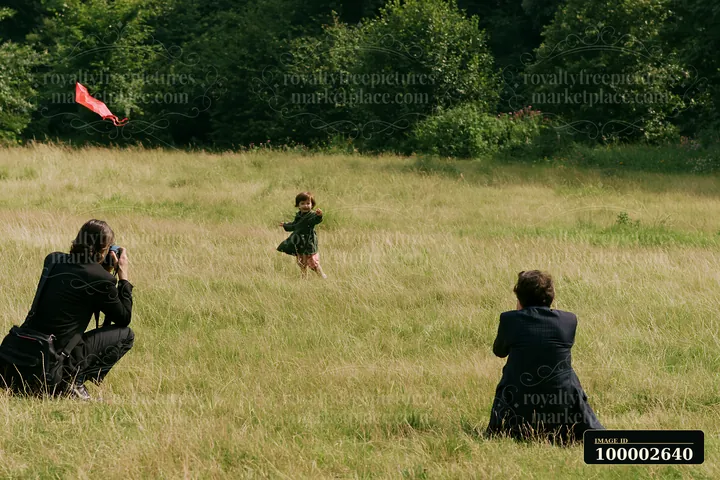 A photographer with camera in meadow takes pictures of a child running and flying a kite.