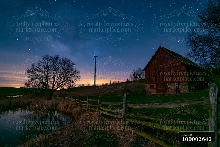 A wind turbine stands near a barn by a river under a starry sky.