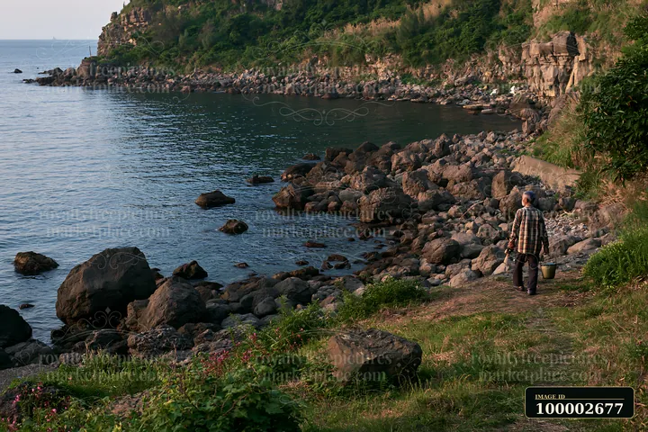 A person walks along a rocky coastline with exposed rocks in the foreground and water in the background.