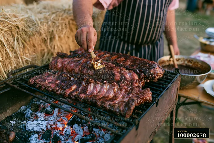 A man brushes barbecue sauce over goat ribs on a grill during a lively countryside festival.