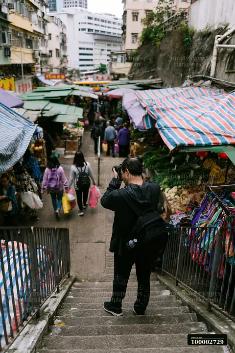 A street photographer taking pictures in an urban market stands on steps overlooking stalls and shoppers.