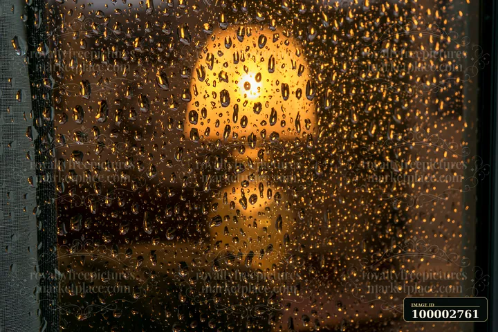 Raindrops on cottage window with warm lamp glow create a cozy atmosphere.
