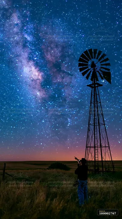 A person using camera under Milky Way stands next to a windmill in a rural area at night.
