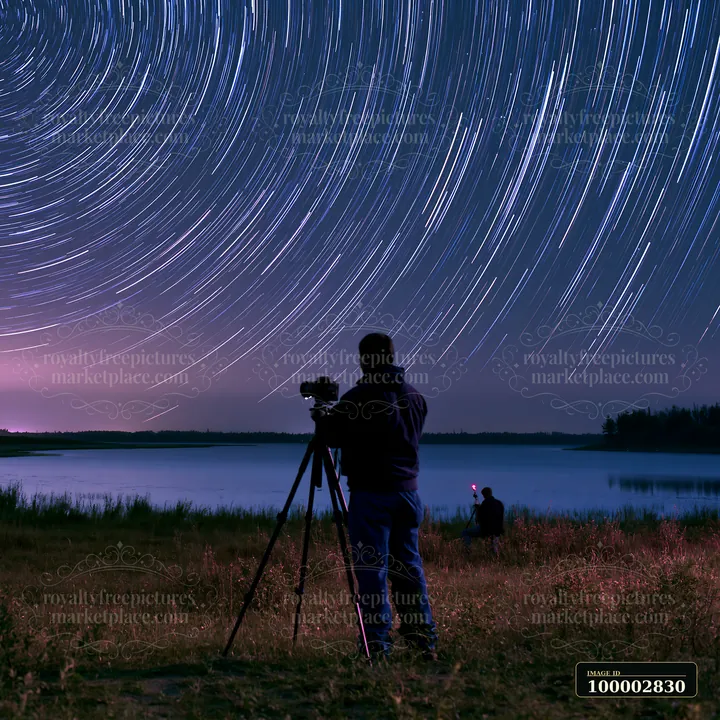 A person stands with a camera on a tripod, capturing star trails above a calm lake surrounded by trees.