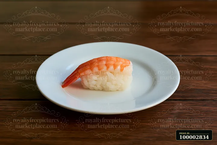 One shrimp sushi piece on a white plate sits on a wooden table.