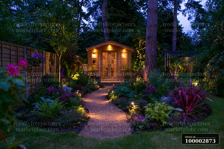 A garden studio stands illuminated at dusk, surrounded by flower beds and pathways lit by soft lights.