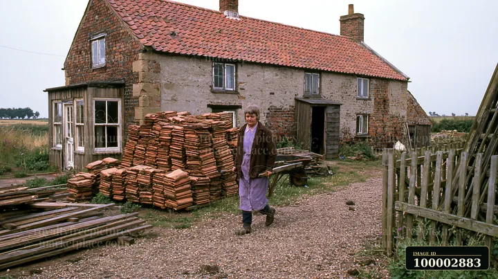 A woman walks near a country house undergoing renovation, with stacks of tiles and wooden planks nearby.