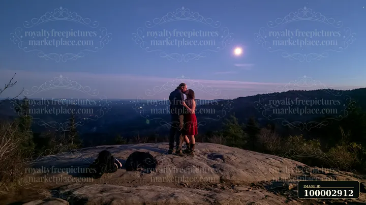 Lovers kissing on a mountain overlook at night under the moonlight, surrounded by trees and mountains, with city lights visible in the distance.