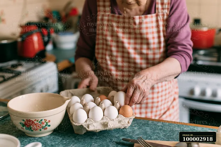 A senior woman with dementia prepares simple food in her kitchen by placing an egg from a carton into a bowl.