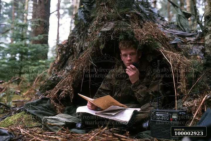 A soldier sits under camouflage netting in a forest, engaged in communication while surrounded by notes and equipment.