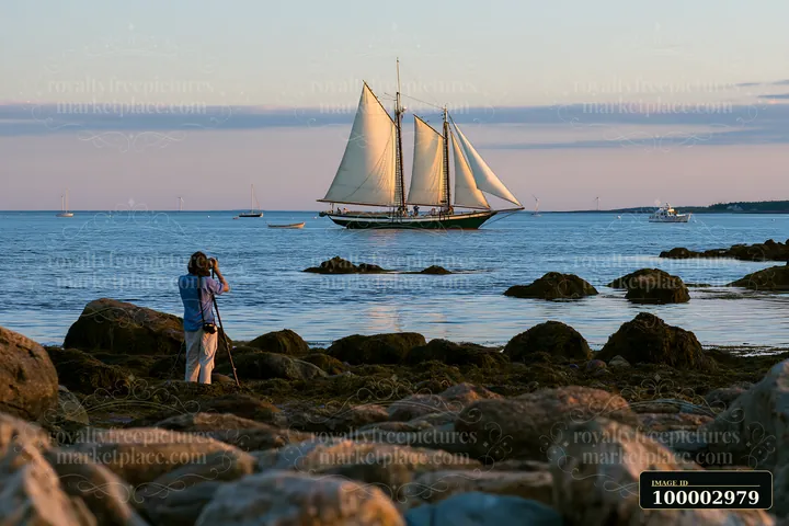 A photographer photographing a sailboat at sea stands on rocky shore.