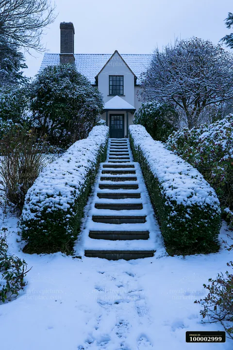 A snowy scene shows a pathway leading to a farmhouse, surrounded by trimmed hedges and trees.