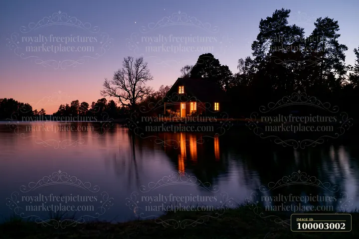 A holiday home under stars stands near a still lake with lights glowing from the windows.