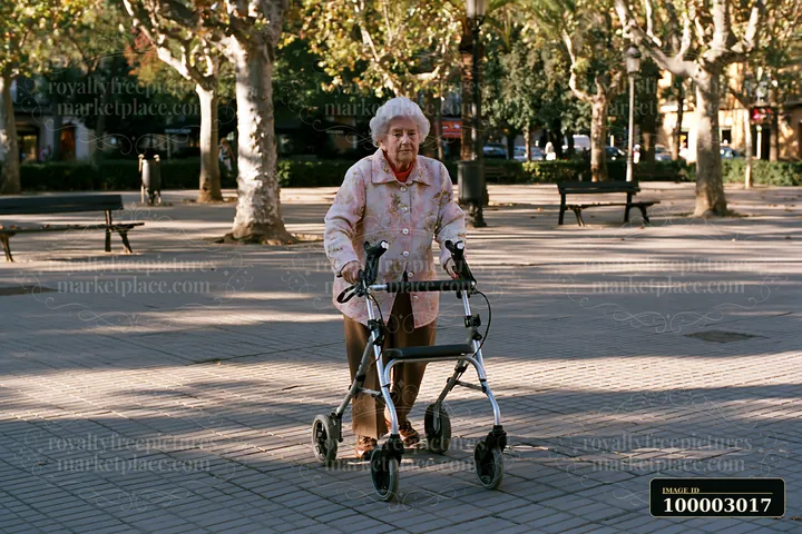 Elderly woman with Parkinson's disease walking outdoors in a park setting, using a walker for support.