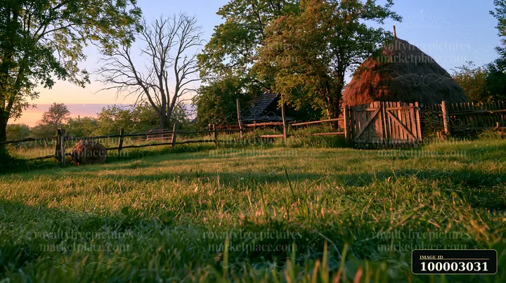 A traditional haystack stands in a meadow at sunset, surrounded by trees and a wooden fence.