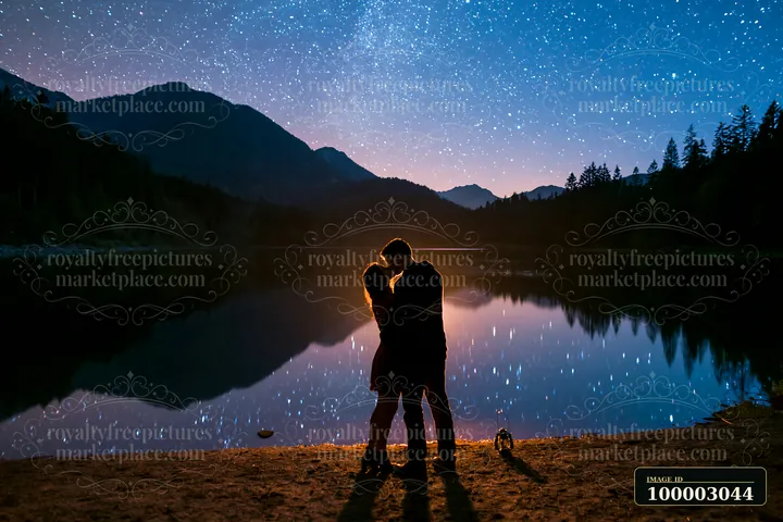 A couple is kissing under stars beside a lake at night, surrounded by mountains and trees.