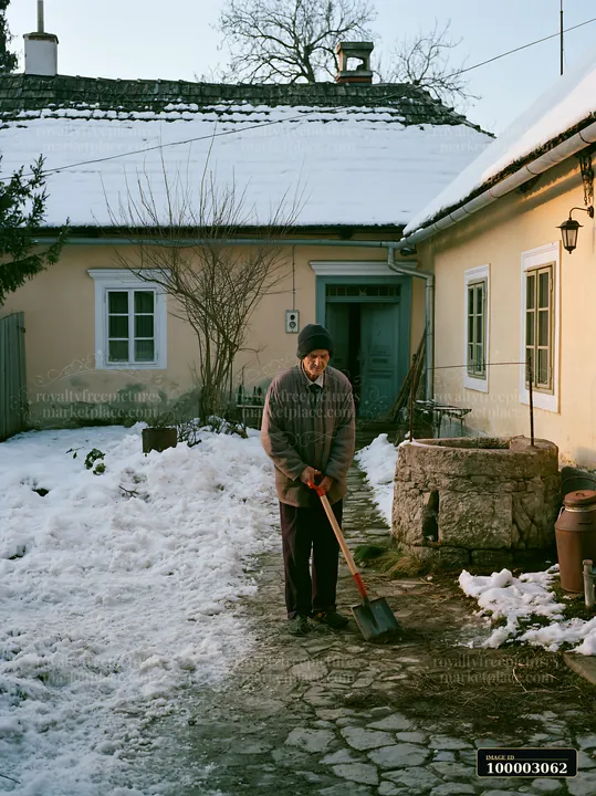 A person shovels snow in a farmhouse courtyard with well and buildings, surrounded by winter scenery.