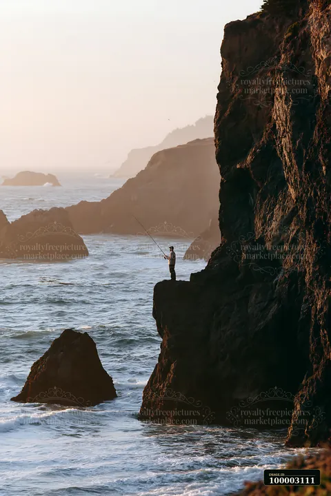 A person stands on a rock near the ocean, fishing along the coastline beneath dramatic cliffs during sunset.