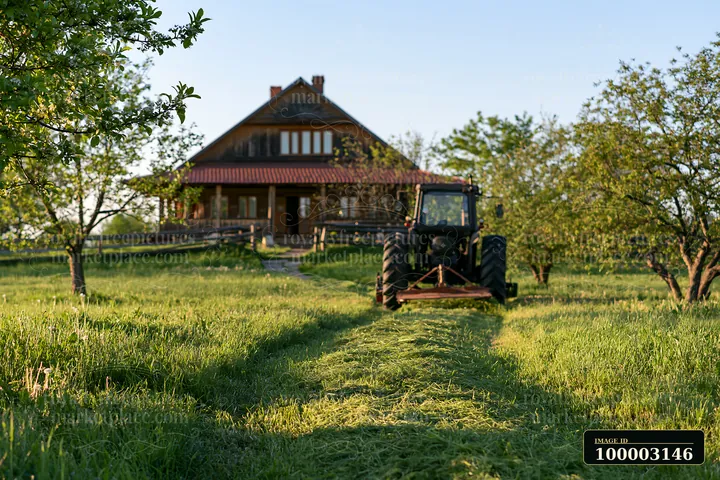 A tractor cuts grass in a meadow leading to a wooden house surrounded by trees.