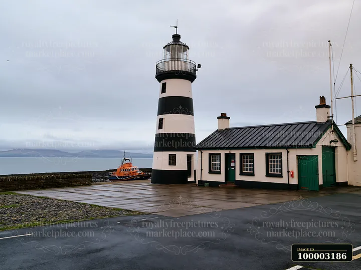 A lighthouse with black and white stripes stands by the sea, next to a small building and an orange boat.
