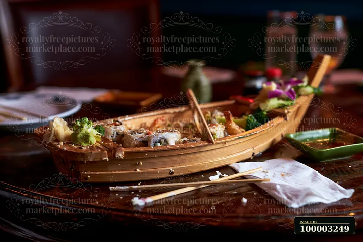 A wooden boat platter features a variety of sushi and garnishes alongside soy sauce in a restaurant setting.