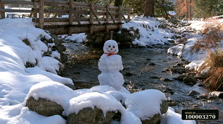 A snowman near a wooden bridge stands by a flowing stream surrounded by snow-covered rocks.