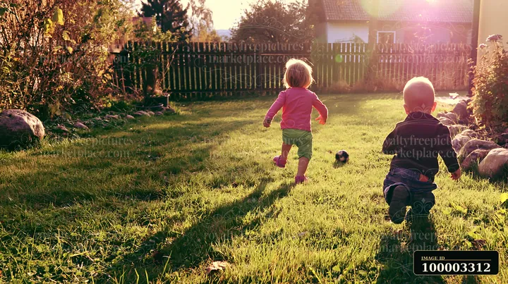 Two young children play in a farmhouse garden, running on the grass as the sun sets behind them.
