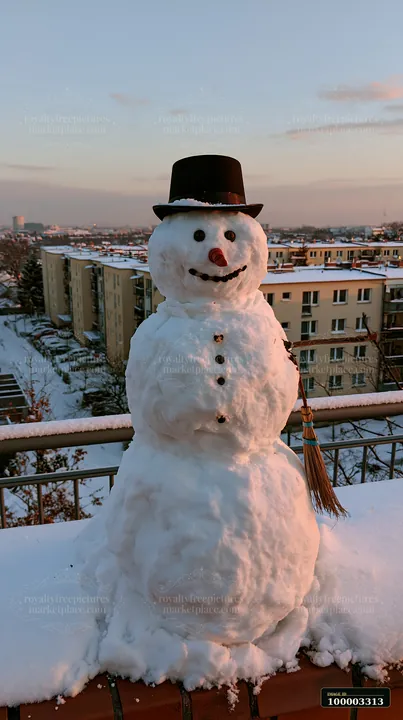 A snowman with a broomstick arm stands on a balcony with a view of apartment buildings.