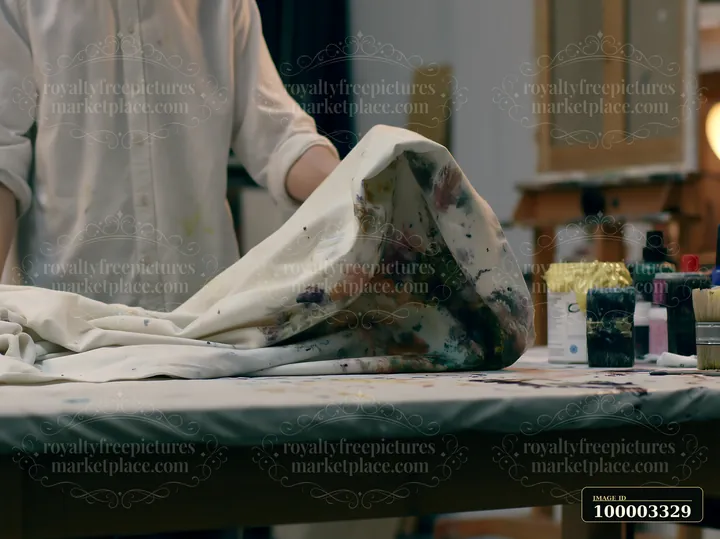 An artist works at a table covered with a splattered artist canvas textile and various painting supplies.