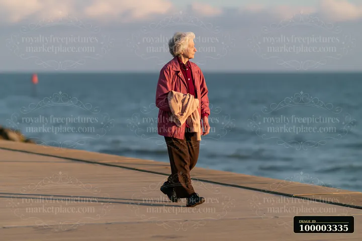 An elderly woman with Parkinson's disease walking outdoors along a pathway near the water.