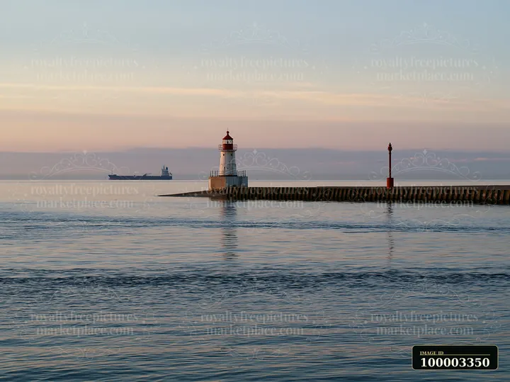 A lighthouse stands on the edge of a pier while a cargo ship can be seen in the distance across the calm water during sunset.