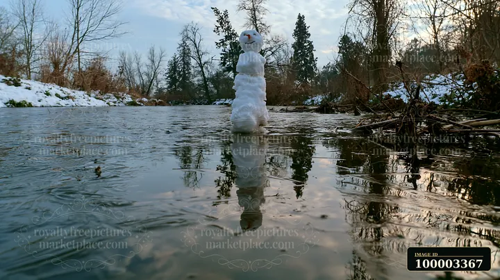 A snowman stands on the edge of a frozen pond, its reflection visible on the icy surface.