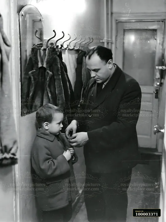 A man assists his young son in putting on a coat in a hallway of a Soviet apartment.