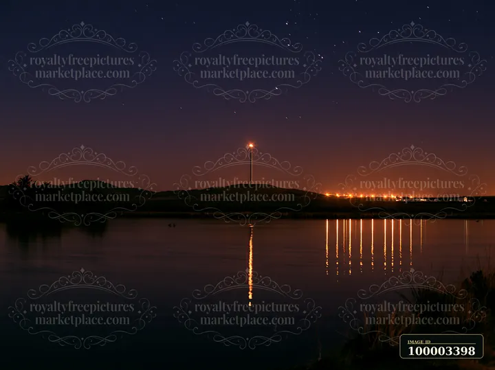 Stars fill the night sky above a rural wind turbine near a calm lake.