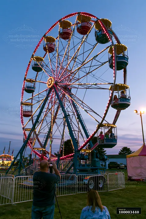 Families enjoy a fair at dusk, with parents photographing children on rides.