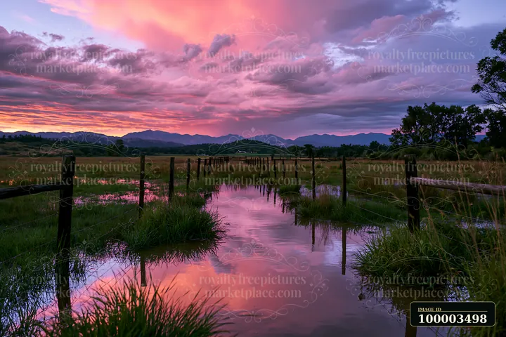 The sky shows a thunderstorm over the mountain range as colorful clouds reflect on a calm body of water.