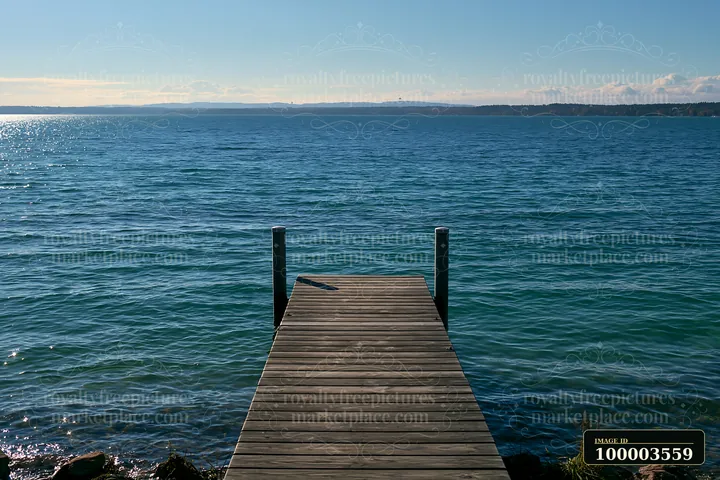 A wooden dock extends over sunlit turquoise water, surrounded by smooth rocks.