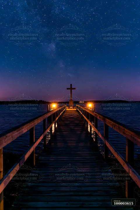 A wooden pier leads to a war memorial cross silhouetted against stars in the night sky.