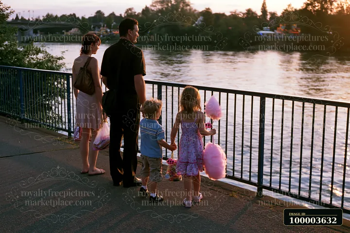 Family walking beside a river at sunset, with children carrying cotton candy and fair prizes.