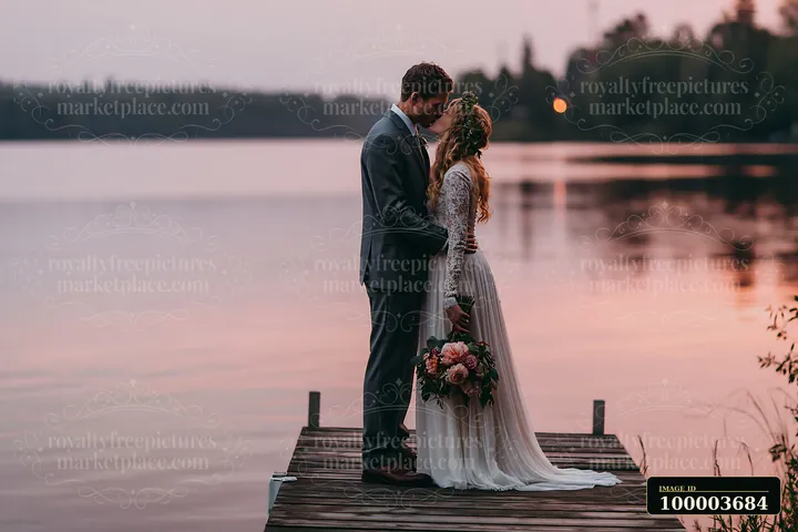 A bride and groom kiss while standing on a wooden dock at sunset near a lake.