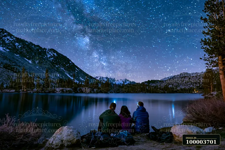 Three hikers sit beside a mountain lake beneath a star-filled sky and snow-dusted peaks.