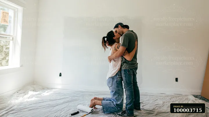 Couple kisses while standing on a covered bedroom floor during a bright home renovation.
