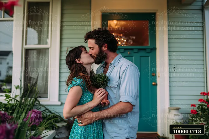 A couple kisses on the porch of a home while holding a bouquet of flowers.